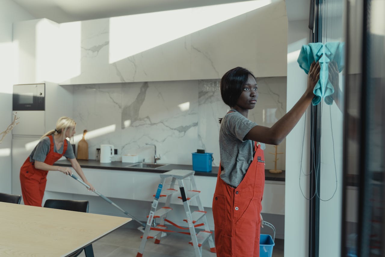 about-02 Two women cleaning a modern kitchen, wiping glass surfaces and mopping the floor.