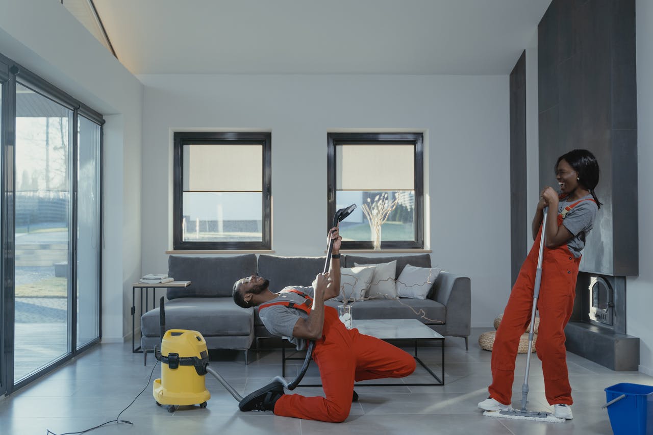 hero-img-01 Fun and engaging cleaning session with a man and woman in overalls using cleaning tools in a modern living room.
